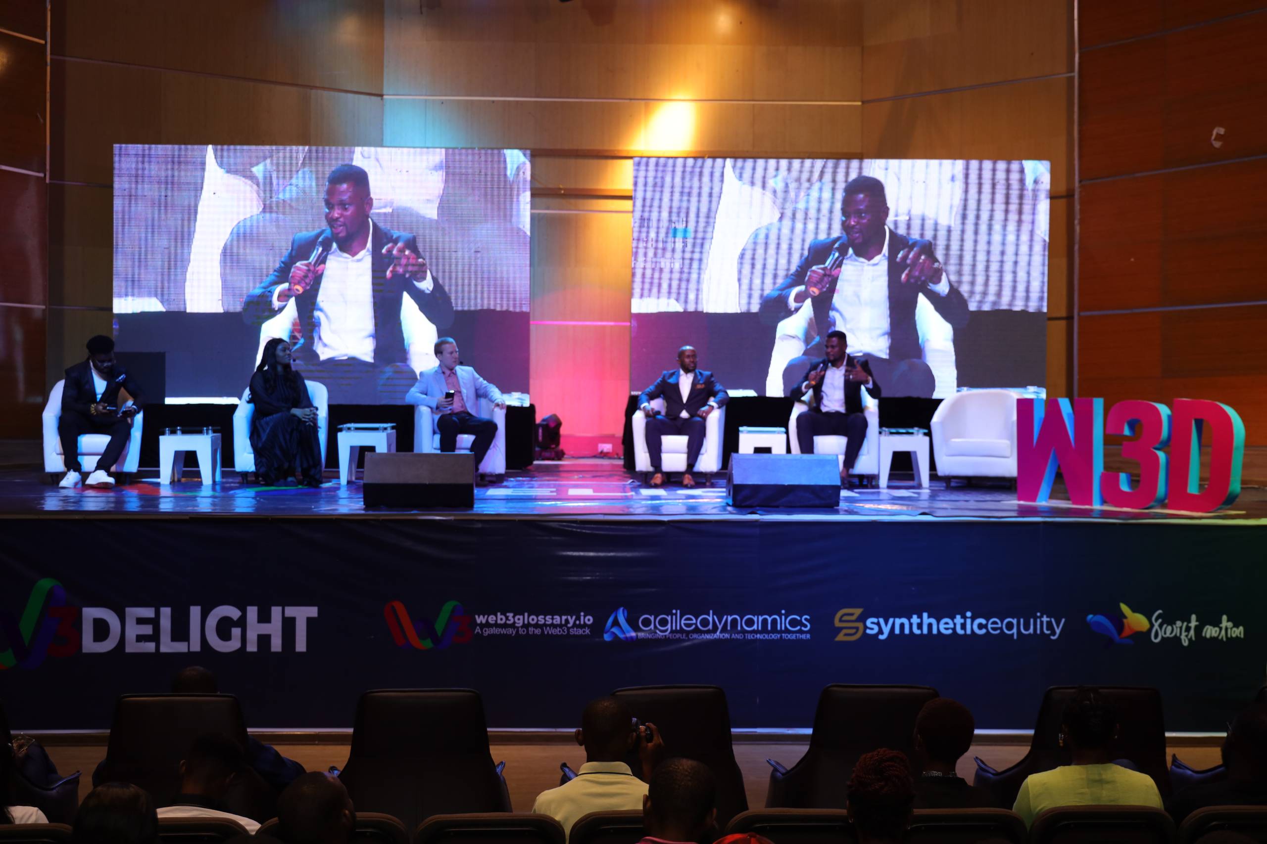 A conference on the Glover Memorial Hall stage, twin LED screens flanking the panel.