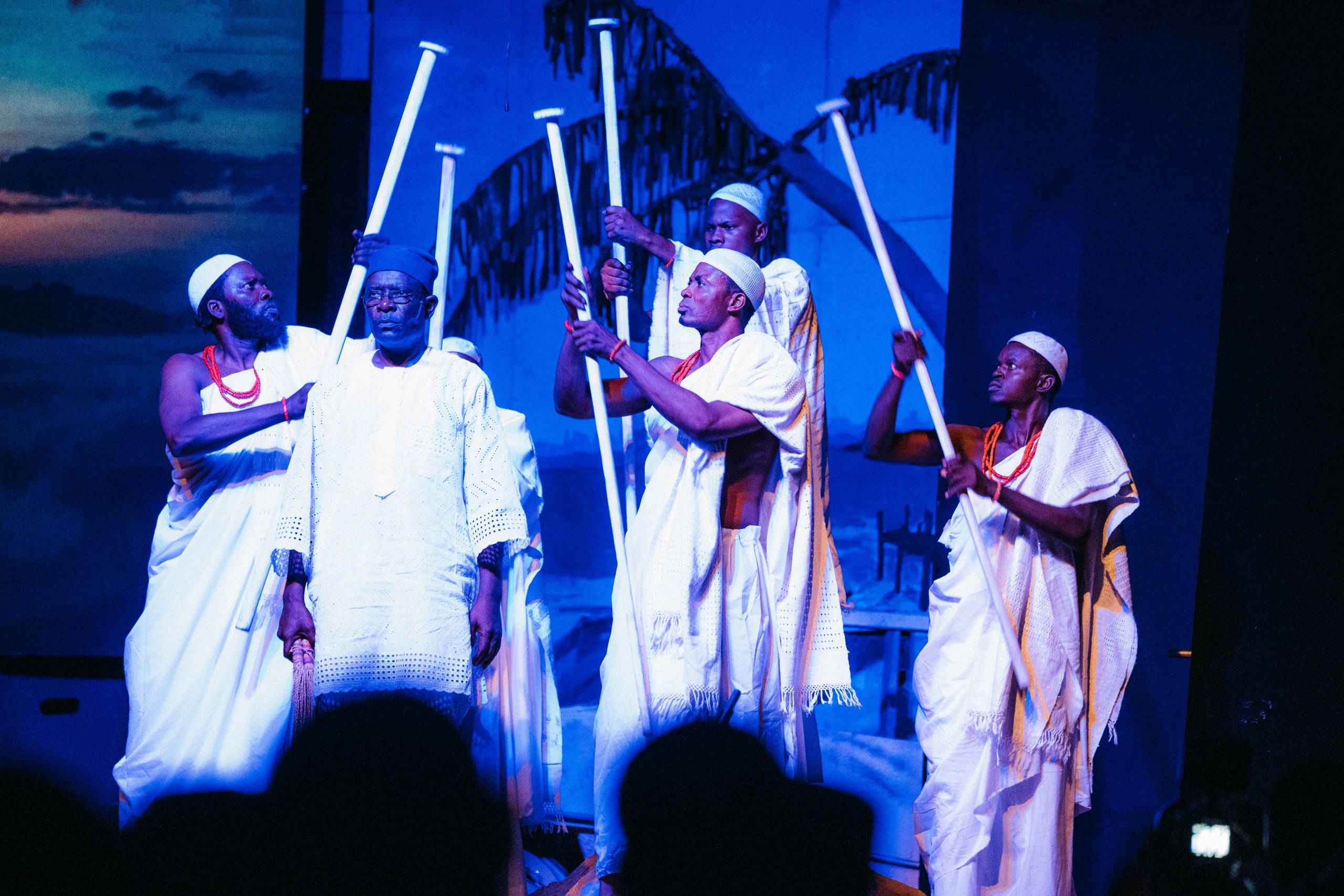 Performers in white agbada raise carved staffs on the stage of Glover Memorial Hall, the audience in silhouette in the foreground.